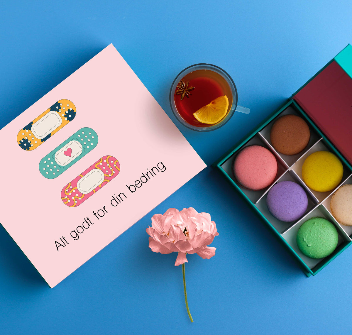 En åpen eske med fargerike makroner ved siden av en rosa gaveeske med tre illustrasjoner av fargerike plaster og teksten &#39;Alt godt for din bedring&#39;. En kopp te og en rosa blomst ligger på en blå bakgrunn.