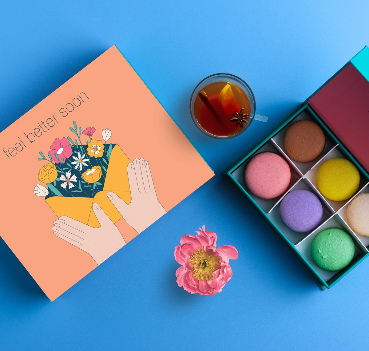 En åpen eske med fargerike makroner ved siden av en oransje gaveeske med teksten &#39;feel better soon&#39; og en illustrasjon av hender som holder en blomsterbukett i en konvolutt. En kopp te og en rosa blomst ligger på en blå bakgrunn.