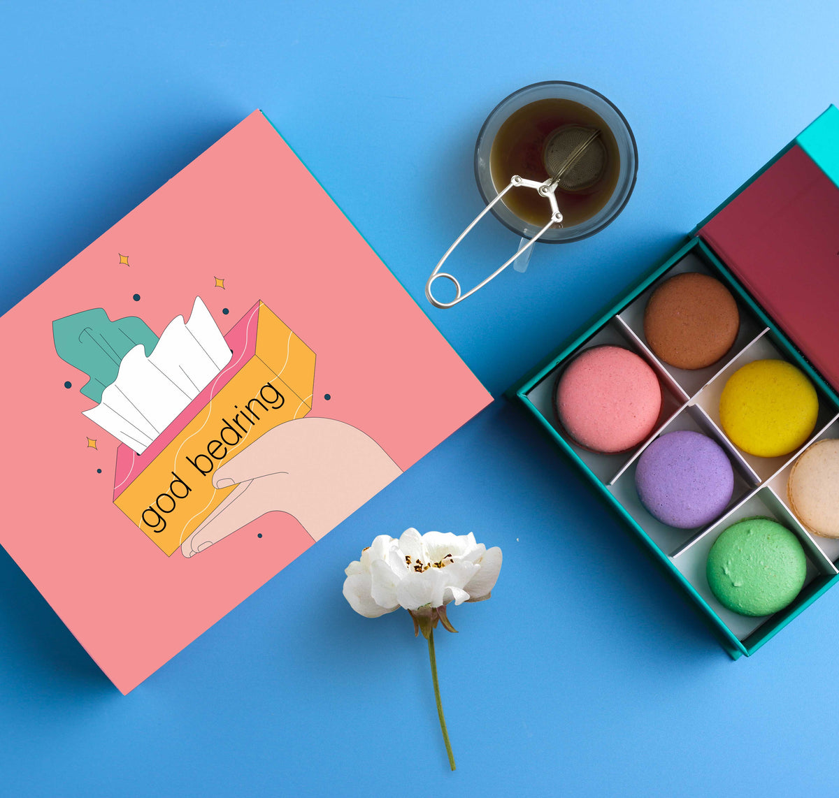 En åpen eske med fargerike makroner ved siden av en rosa gaveeske med tre illustrasjoner av fargerike plaster og teksten &#39;Alt godt for din bedring&#39;. En kopp te og en rosa blomst ligger på en blå bakgrunn.
