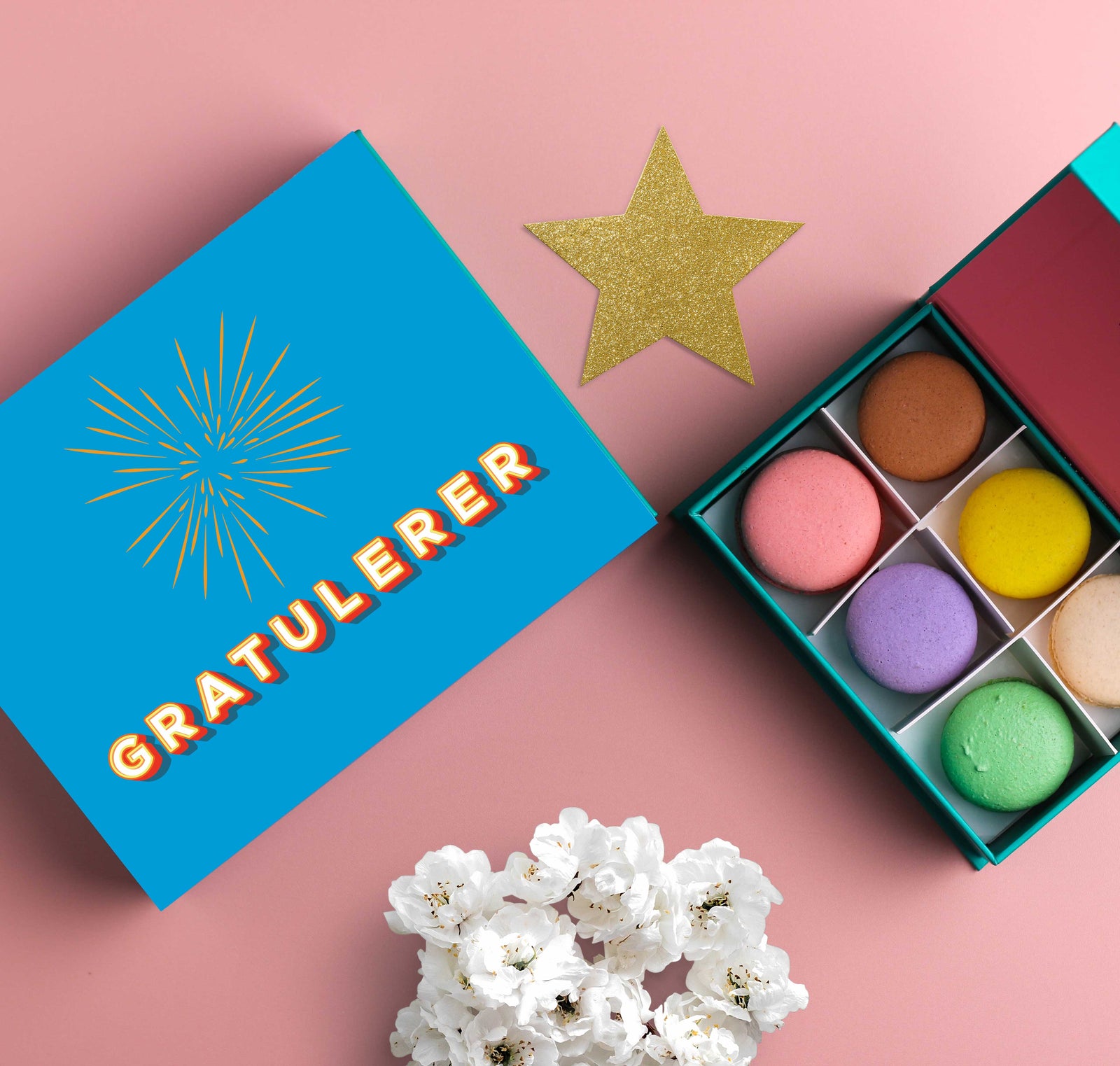 En åpen eske med fargerike makroner ved siden av en blå gaveeske med teksten 'Gratulerer' og en illustrasjon av et fyrverkeri. En gullglitterstjerne og hvite blomster ligger på en rosa bakgrunn.